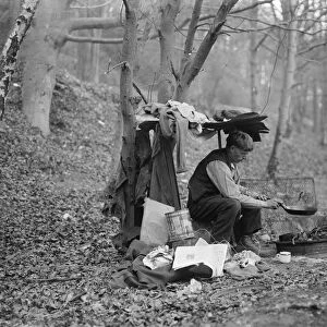 Tramp at home in woods cooking food over a camp fire. 1935