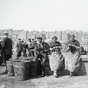 Female herring fishers, knitting on the dock, in front of empty buckets waiting for the return of fishing, in Scarborough (Yorkshire, England), 1910 (b/w photo)